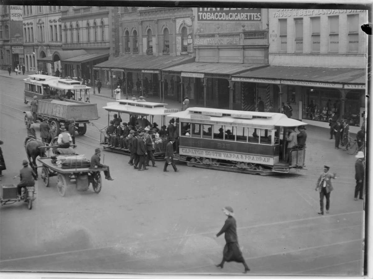 Truck towing tram in procession along street and view of shop fronts such as Reaby Taylor Pty Ltd and the Melbourne Cafe, Prahran (1902–20)