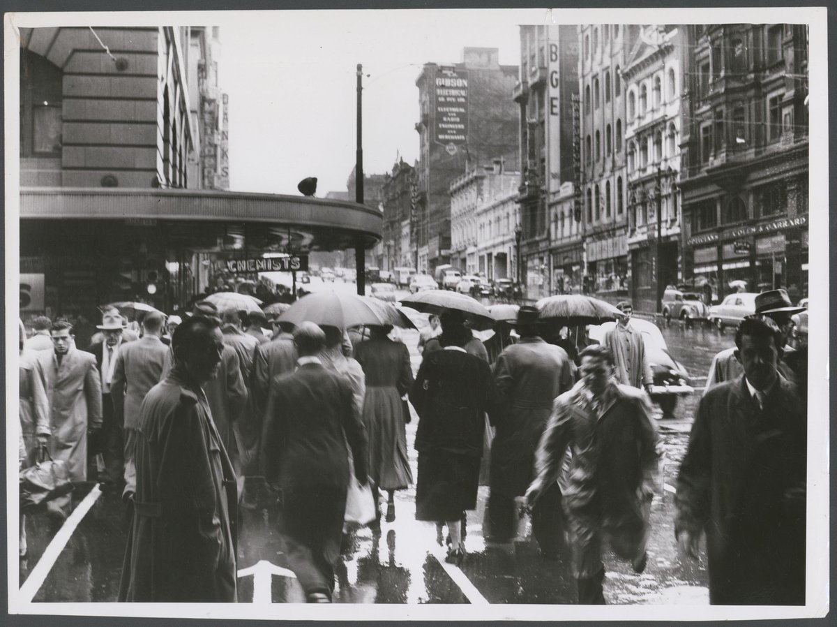 Hurrying across Elizabeth St. on the south side of Bourke St. through the rain today, Melbourne City (1954)