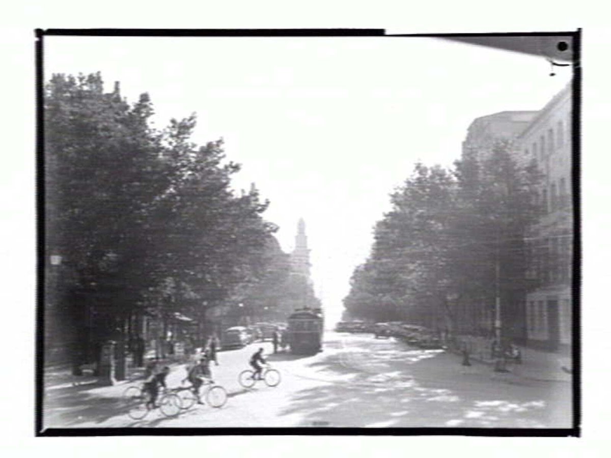 View down Collins Street from Spring Street, with bicyclists crossing the street, Melbourne City (1936–39)