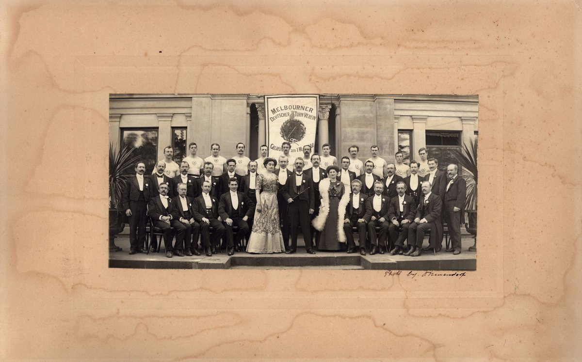 Group of men and two women standing before banner reading: Melbourner Deutscher Turn Verein (1886–98)