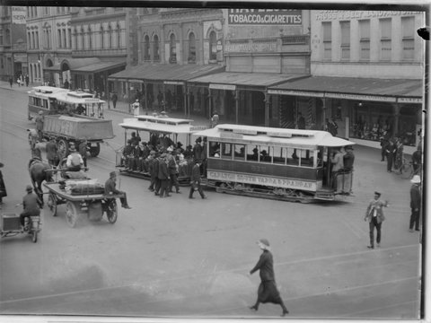 Truck towing tram in procession along street and view of shop fronts such as Reaby Taylor Pty Ltd and the Melbourne Cafe, 1902–20