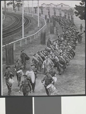 Troops marching behind drummers and officer along side tram or train tracks, Royal Park, Melbourne, c. 1940