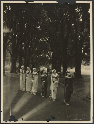 Six women in single file wearing "Grecian costume" in the Fitzroy Gardens, c. 1920
