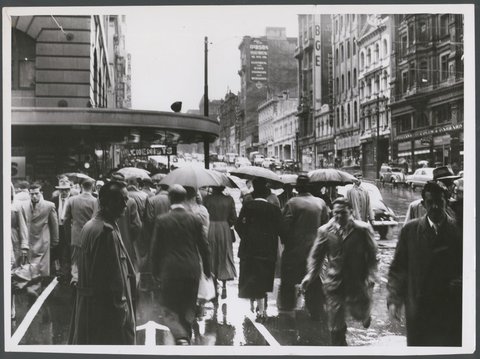 Hurrying across Elizabeth St. on the south side of Bourke St. through the rain today, 1954