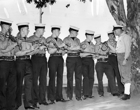Bends and hitches occupy the hands, eyes and minds of these National Service trainees under the eagle eye of Chief Petty Officer G.L. Davies, of Melbourne, during their seamanship course, c. 1951