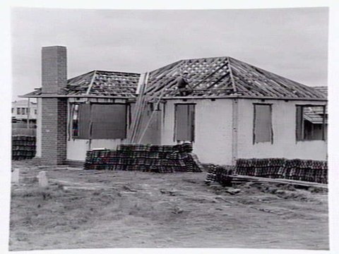 Housing Commission estate homes under construction at Ashburton, showing stumps, roof before tiling and cranes putting up concrete, pre-fabricated walls, c. 1946