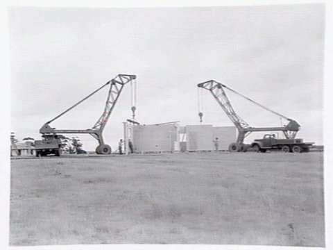 Housing Commission estate homes under construction at Ashburton, showing stumps, roof before tiling and cranes putting up concrete, pre-fabricated walls, c. 1946