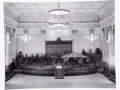 South Melbourne Town Hall, stage area decorated with plants and bark roofed hut, c. 1950