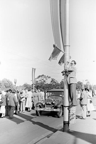 Man standing on top of a rubbish bin and holding on to a lightpole on Princes Bridge, to get a better view over the crowds, in Melbourne during the royal tour of 1954